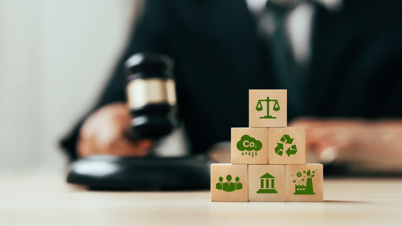 Sechs gestapelte Würfel mit grünen Bildzeichen, die Justiz, Nachhaltigkeit und erneubare Energien darstellen. Im Hintergrund schlägt ein Richter mit seinem Hammer auf dem Tisch.