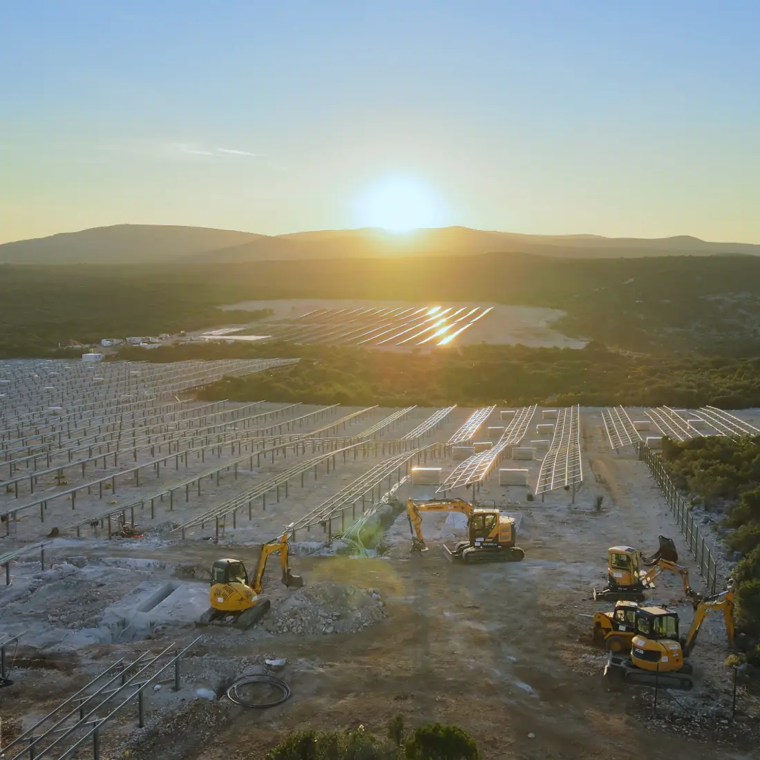 Aerial view over machinery, pilons, and naked module frames at a solar farm construction site, during sunset
