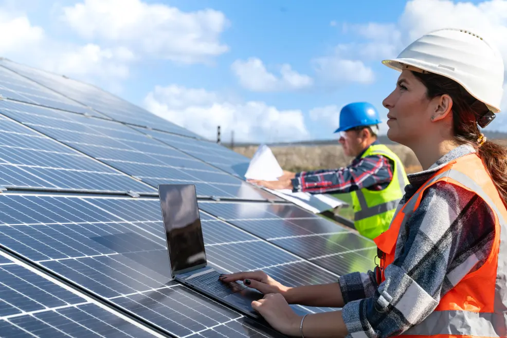 Image of a female solar engineer using her laptop, with the solar module of a utility-scale solar power plant as a table. In the background, another solar engineer extends a paper blueprint over another solar panel.